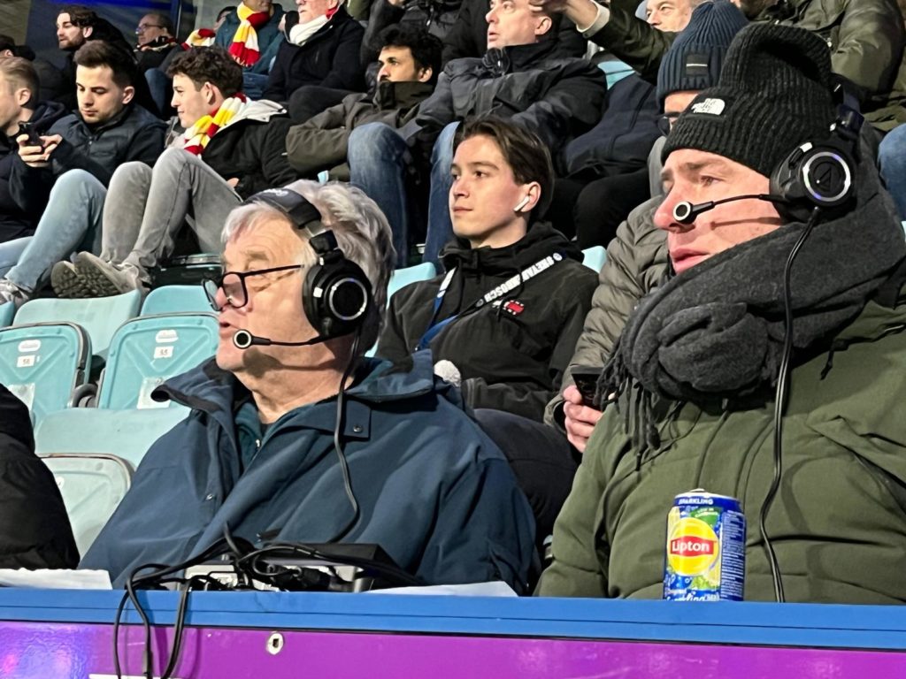 Twee mannen zitten, warm aangekleed, vooraan op de tribune in het stadion, met koptelefoon op en apparatuur om het audioverslag te maken.